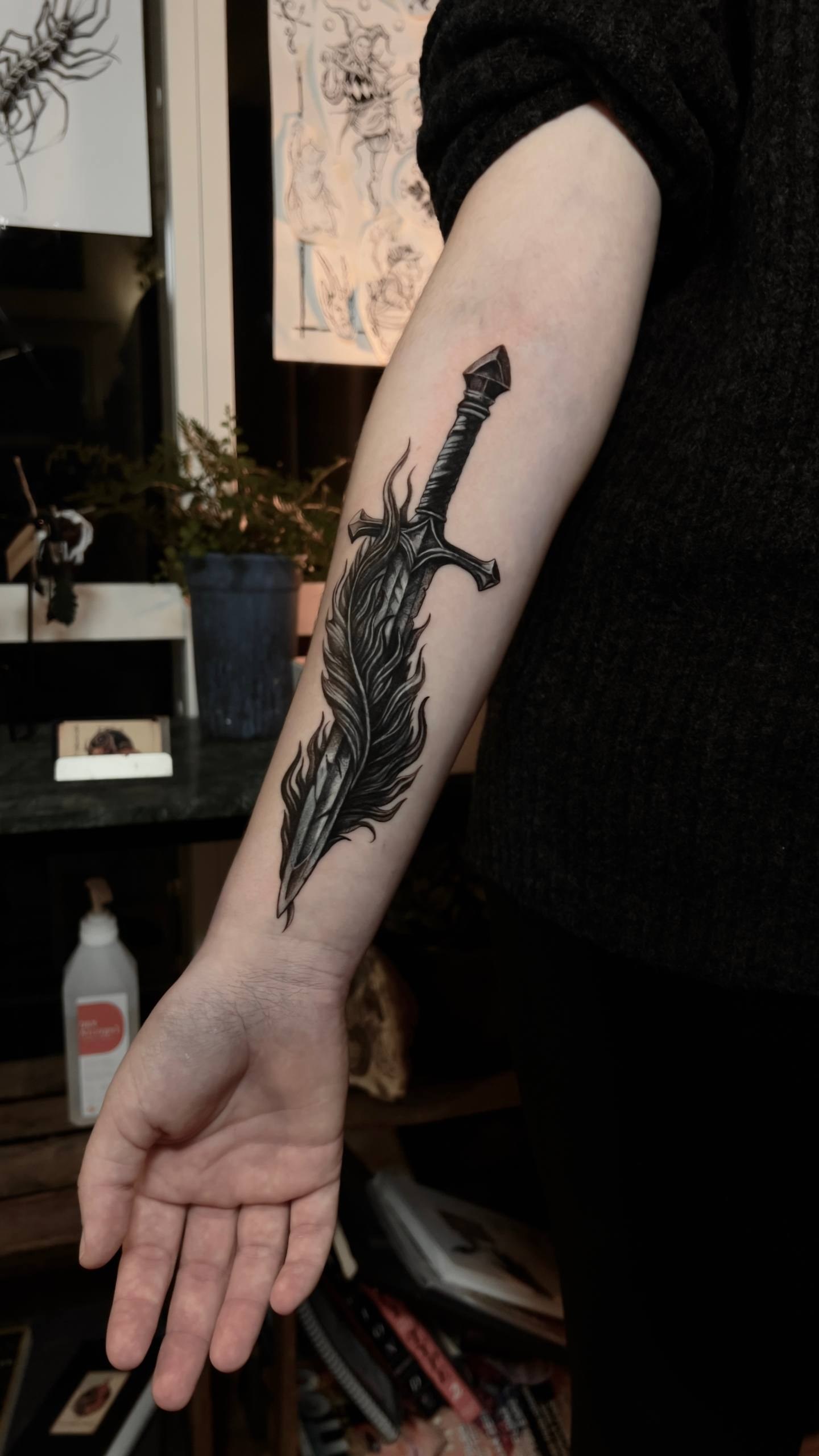 Mörk fantasy tatuering Uppsala — svärd och fjäder i blackwork, underarm